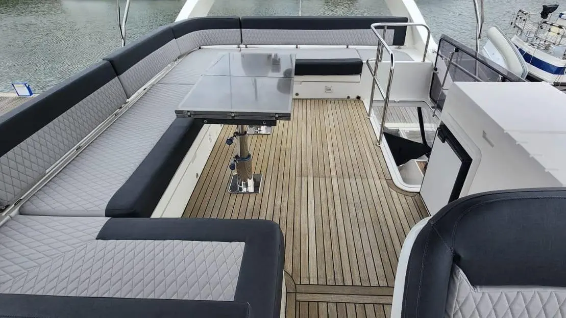 Luxury boat deck featuring grey and black cushioned seating, a glass-top table, and wooden flooring beside moored boats.